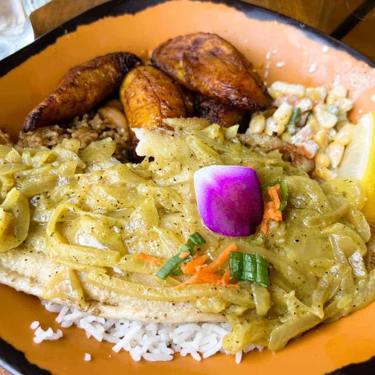 Samaki, a pan flash-fried fish topped with tangy sautéed onions. A fresh corn salad, fried plantains, stewed yellow beans, and your choice of spiced rice pilau or coconut rice accompany the fish. 